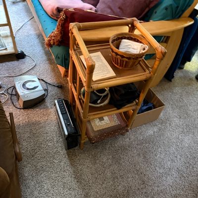 Sale Photo Thumbnail #23: Vintage three-tier bamboo and rattan shelf unit holding books and a woven basket, positioned next to a wooden futon sofa and portable radios.