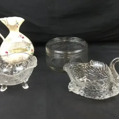 Sale Photo Thumbnail #119: Collection of decorative glassware and porcelain pieces, including an etched footed bowl, a clear glass swan dish, a round covered glass container, and a small floral porcelain pitcher.