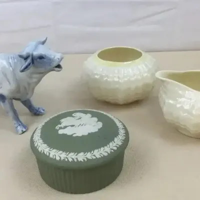 Sale Photo Thumbnail #24: Collection of decorative porcelain and ceramic items, including a green Wedgwood jasperware trinket box, a Delft-style cow creamer, and a pair of iridescent cream glaze vessels.