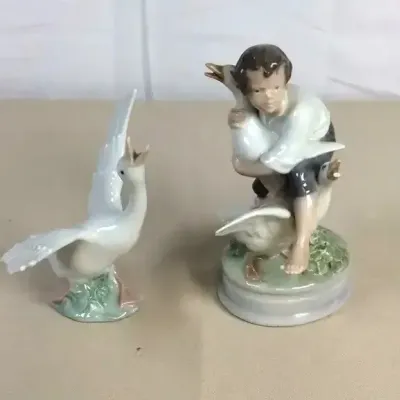 Sale Photo Thumbnail #54: Two porcelain figurines featuring a goose with wings spread and a young boy wrestling with geese, crafted in a soft pastel finish.