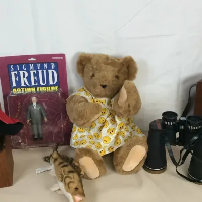 Sale Photo Thumbnail #109: Eclectic assortment of items including a vintage teddy bear in a smiley face dress, a packaged Sigmund Freud action figure, a pair of black binoculars with a leather case, and novelty stuffed figures.