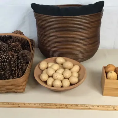 Sale Photo Thumbnail #53: Rustic home decor collection including a wooden basket filled with pine cones, a bowl of faux speckled eggs, a small box with decorative balls, and a large coiled dark wood basket.