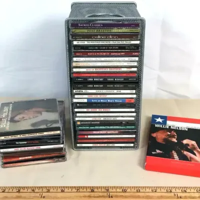 Sale Photo Thumbnail #63: Collection of compact discs including pop, rock, country, and jazz titles, organized in a metal mesh storage rack alongside small stacks.