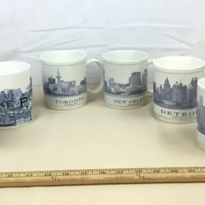Sale Photo Thumbnail #2: Collection of six ceramic souvenir coffee mugs featuring city skylines and regional themes including Texas, Toronto, and Denver.