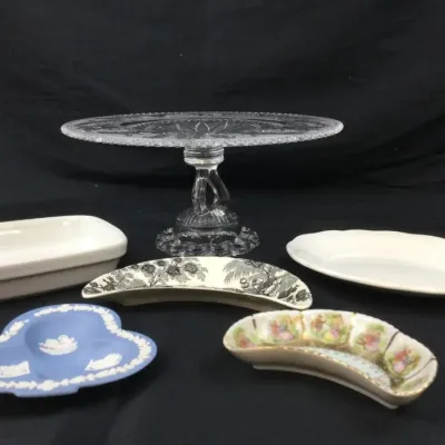 Sale Photo Thumbnail #66: Assortment of vintage tabletop serveware, including a pressed glass pedestal cake stand, a blue Wedgwood Jasperware tricorn dish, crescent bone dishes, and white ceramic trays.