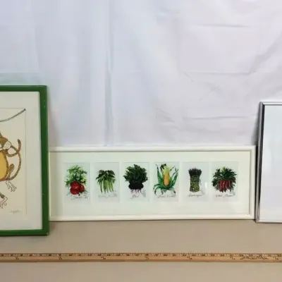 Sale Photo Thumbnail #65: Trio of framed wall art, including a whimsical monkey illustration in a green frame, a horizontal botanical vegetable print, and a modern portrait print in a metal frame.