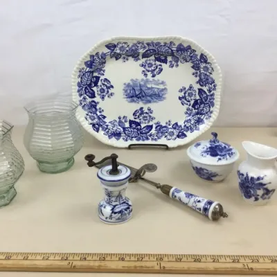 Sale Photo Thumbnail #70: Collection of blue and white tableware including an oval serving platter with a nautical scene, sugar bowl, creamer, mill, and server, displayed alongside two clear glass hurricane-style vases.