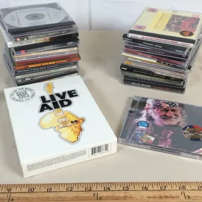 Sale Photo Thumbnail #52: Collection of compact discs and box sets, featuring a Live Aid 1985 DVD set, a Willie Nelson CD/DVD, and two stacks of various music CDs.