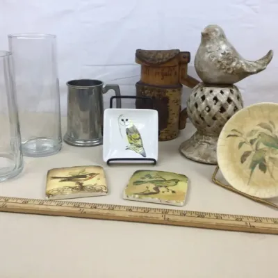Sale Photo Thumbnail #84: Assortment of decorative items featuring bird-themed ceramic coasters and small plates, clear glass cylinder vases, a pewter mug, and a carved wooden vessel.