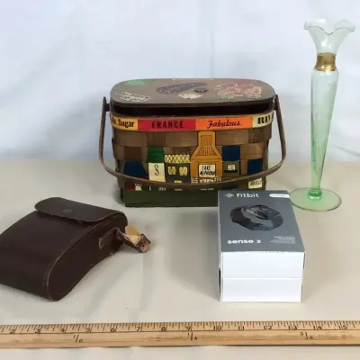 Sale Photo Thumbnail #40: Assortment of small goods including a vintage Caro Nan style hand-painted bucket handbag, a green uranium glass vase, a boxed Fitbit Sense 2, and a brown leather case.