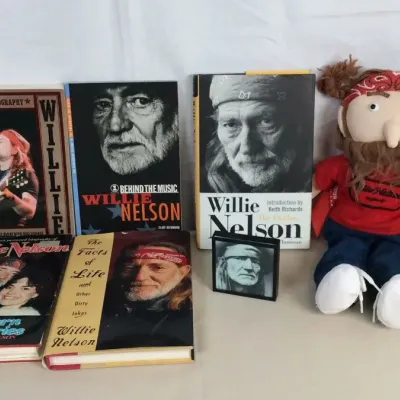 Sale Photo Thumbnail #118: Collection of Willie Nelson memorabilia, including five books, a small framed portrait, and a plush Willie Nelson doll wearing a red bandana.