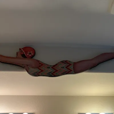 Sale Photo Thumbnail #121: Art Deco style wooden cutout wall art depicting a female swimmer in a patterned bathing suit and red swim cap, mounted above vanity lighting.