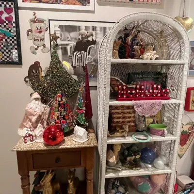 Sale Photo Thumbnail #71: A collection of Christmas decor displayed on a white wicker shelf and a small wooden end table, surrounded by vintage-style wall art.