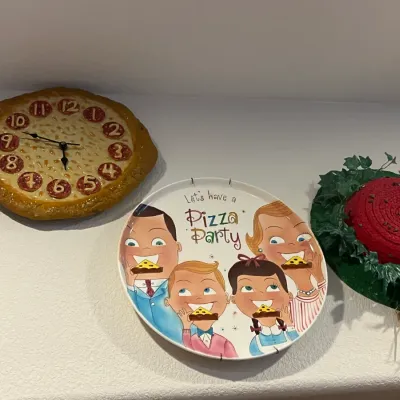 Sale Photo Thumbnail #61: Novelty wall decor featuring a pepperoni pizza wall clock, a retro "Let's Have a Pizza Party" illustrated ceramic platter, and a watermelon-themed straw hat hanging.