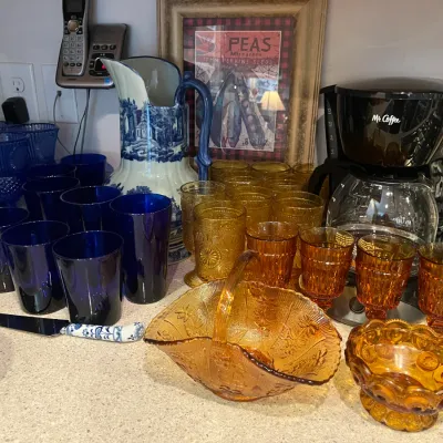 Sale Photo Thumbnail #27: Collection of vintage glassware featuring amber depression glass pieces, cobalt blue goblets and tumblers, a blue and white ceramic pitcher, and a Mr. Coffee maker on a countertop.