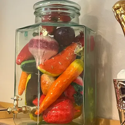 Sale Photo Thumbnail #33: Large clear glass beverage dispenser with a brass spigot, filled with colorful decorative faux fruits and vegetables.
