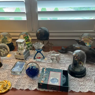 Sale Photo Thumbnail #206: Collection of decorative glass paperweights, snow globes, and figurines displayed on a white lace runner, including a dark shark sculpture and a boxed Spirit of St. Louis paperweight.