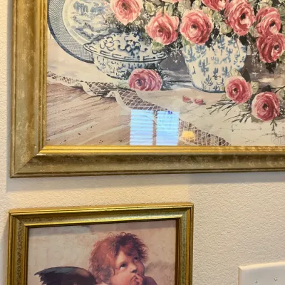 Sale Photo Thumbnail #177: Two framed wall art prints in antiqued gold-toned frames, including a floral still life featuring pink roses and blue china, and a smaller print depicting Raphael's cherub.