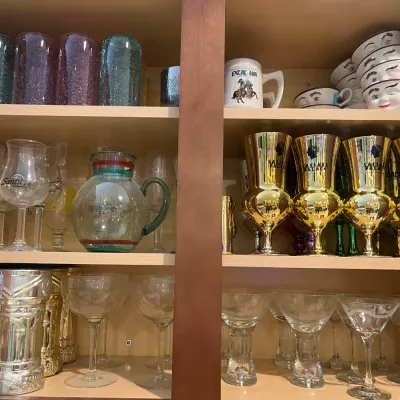 Sale Photo Thumbnail #22: Wooden cabinet filled with novelty drinkware and glassware, including Baileys winking face mugs, metallic Las Vegas souvenir goblets, and colorful tumblers.