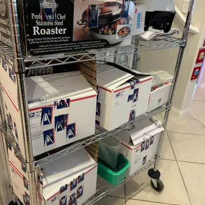 Sale Photo Thumbnail #59: Chrome wire rolling baker's rack loaded with boxed kitchen items, including a Professional Chef stainless steel roaster, small appliances, and USPS Priority Mail shipping boxes.