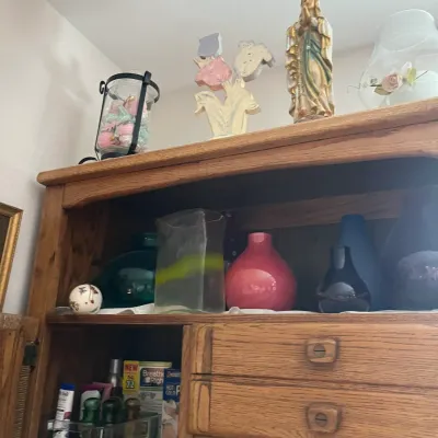 Sale Photo Thumbnail #195: A wooden cabinet and hutch displays various decorative items, including colorful glass vases, a religious statue, and a lantern.