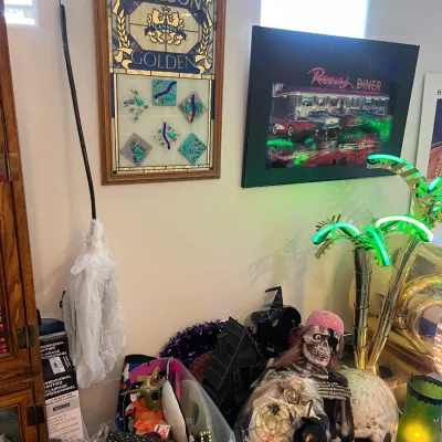 Sale Photo Thumbnail #158: A collection of Halloween decorations, framed wall art, including a Molson Golden stained-glass sign and a Rosie's Diner print, alongside a neon palm tree floor lamp.