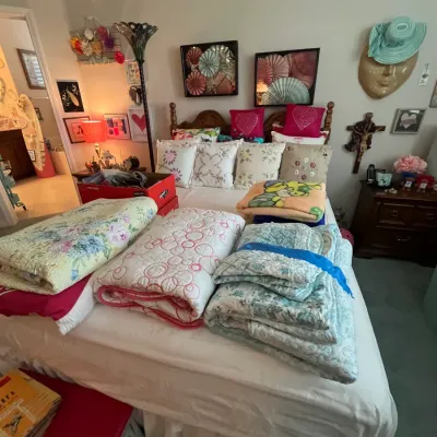 Sale Photo Thumbnail #112: Bedroom set up featuring folded quilts, comforters, and throw pillows displayed on a bed frame, along with wall art, a nightstand, and decorative accessories.