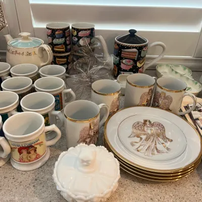 Sale Photo Thumbnail #18: Collection of tabletop ceramics and glassware including vintage advertising mugs, angel-patterned Christmas china with gold trim, teapot sets, and milk glass dishware.
