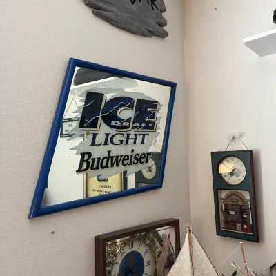 Sale Photo Thumbnail #204: Framed vintage Ice Draft Light Budweiser bar mirror in a blue wooden frame, displayed alongside wall decor including a rustic Tiki Bar sign and clocks.