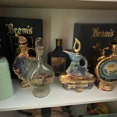 Sale Photo Thumbnail #78: Collection of vintage Jim Beam decanters, including ornate gilded ceramic and glass bottles, displayed on a white shelf alongside original presentation boxes.