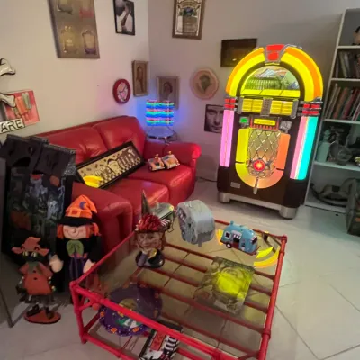 Sale Photo Thumbnail #73: Retro entertainment room featuring a illuminated vintage-style Wurlitzer jukebox, red leather sofa, and red bamboo glass-top coffee table surrounded by various collectibles and novelty decor.