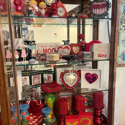 Sale Photo Thumbnail #160: A wooden curio cabinet filled with Valentine's Day-themed decor, including red pillar candles, heart-shaped frames, small statues, clocks, and decorative signs.