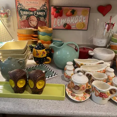 Sale Photo Thumbnail #20: Collection of vintage kitchenware and decor items, including colorful Fiesta-style pitchers and bowls, a pair of carved owl salt and pepper shakers, fruit-patterned tea sets, and decorative wall art.