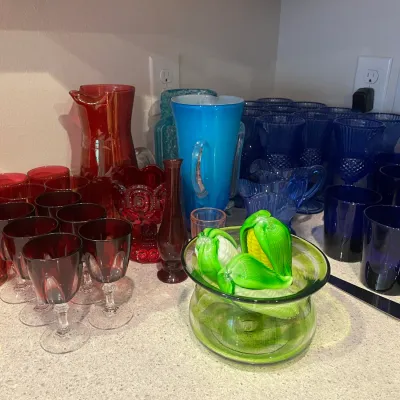 Sale Photo Thumbnail #25: Collection of vintage colorful glassware including ruby red goblets and pitchers, cobalt blue tumblers, a light blue cased glass pitcher, and glass corn on the cob figurines in a green bowl.