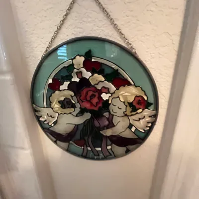 Sale Photo Thumbnail #132: Circular leaded faux stained glass suncatcher featuring two cherubs with a bouquet of flowers, complete with a silver-tone hanging chain.