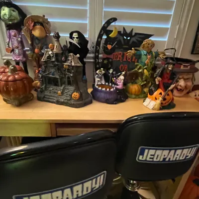 Sale Photo Thumbnail #75: Pair of black vinyl Jeopardy! branded swivel barstools positioned in front of a wooden table displayed with various Halloween decorations.