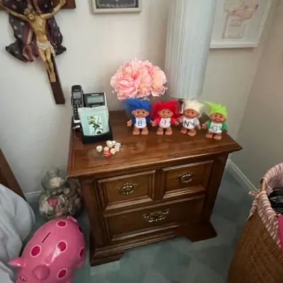 Sale Photo Thumbnail #110: Traditional wooden nightstand featuring vintage troll dolls spelling "BING", a tall cylinder lamp, a pink piggy bank, and various wall decor.