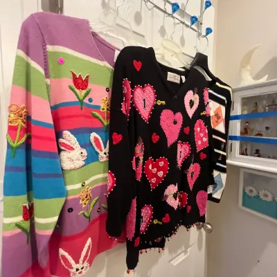 Sale Photo Thumbnail #122: Collection of vintage novelty knit sweaters hanging on a rack, featuring a pastel spring bunny and flower motif and a black heart and key design.