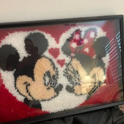 Sale Photo Thumbnail #237: Framed latch hook yarn artwork depicting Mickey and Minnie Mouse kissing, set under glass in a black frame.