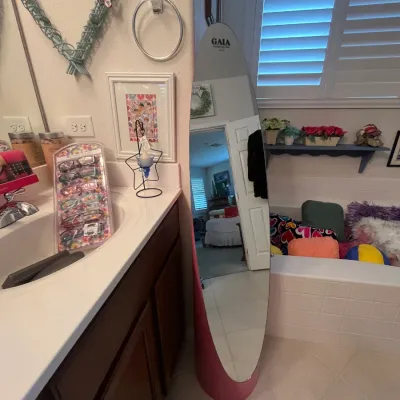 Sale Photo Thumbnail #129: Gaia brand surf-shaped standing full-length mirror with a pink cylindrical base, placed alongside a display of colorful reading glasses on a bathroom counter.