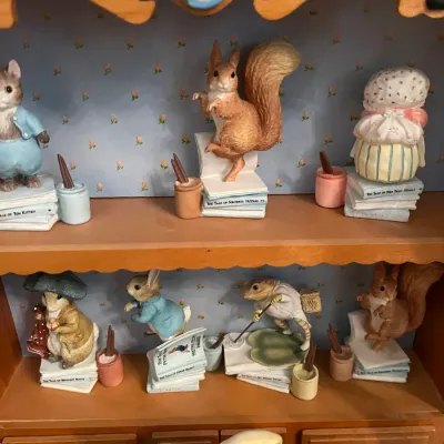 Sale Photo Thumbnail #170: Collection of Beatrix Potter storybook character figurines displayed inside a wooden hutch shelf. Features characters like Peter Rabbit, Squirrel Nutkin, and Mrs. Tiggy-Winkle standing atop stack-of-books bases.