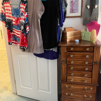 Sale Photo Thumbnail #192: Standing wooden jewelry armoire with brass pulls next to a door hung with women's t-shirts and tops.
