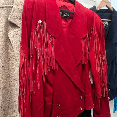 Sale Photo Thumbnail #134: Vintage red suede Western-style jacket with fringe trim and silver-tone concho accents hanging on a clothing rack.