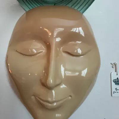 Sale Photo Thumbnail #111: Glossy beige ceramic wall mask sculpture with closed eyes, topped with a green brimmed sun hat.