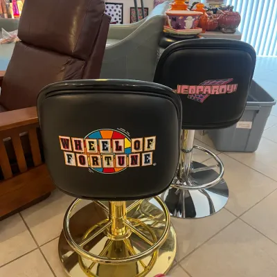 Sale Photo Thumbnail #4: Pair of swivel bar stools featuring embroidered Wheel of Fortune and Jeopardy! logos on black vinyl seats, with metallic pedestals and ring footrests.