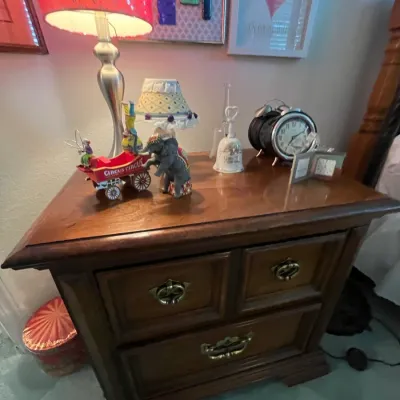 Sale Photo Thumbnail #118: Traditional wooden nightstand with brass drawer pulls, holding table lamps, decorative figurines, and a vintage-style alarm clock.