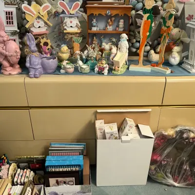 Sale Photo Thumbnail #171: Collection of Easter and spring-themed decor, including rabbit figurines, miniature wooden houses, decorative signs, and boxed items displayed on a long beige cabinet.