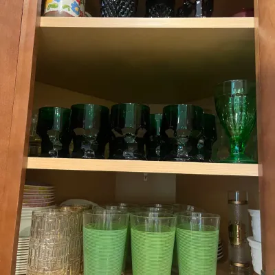 Sale Photo Thumbnail #30: Collection of vintage glassware organized on cabinet shelves, including green striped tumblers, dark green stemware, textured clear glasses, and dark pressed glass pieces.