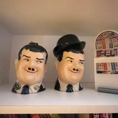 Sale Photo Thumbnail #77: Pair of Laurel and Hardy ceramic character decanters alongside a miniature slot machine toy on a white shelf.