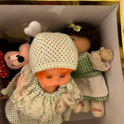 Sale Photo Thumbnail #180: Collection of vintage dolls and plush toys stored in a gold-rimmed box, including a small Cabbage Patch-style doll, a polka-dot dressed baby doll, and a Minnie Mouse plush.